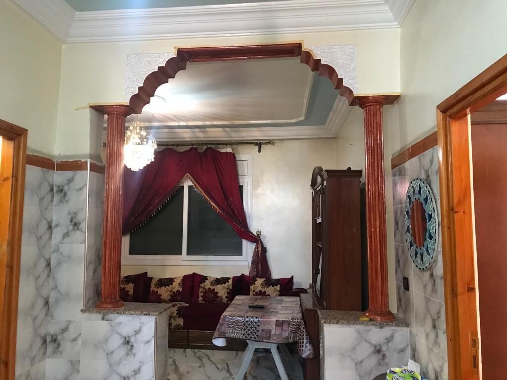 Una habitación con piso de mármol, ventana de madera con cortinas rojas, una mesita y un sofá con cojines estampados.