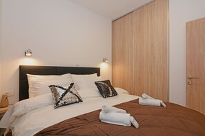 A neatly made bed with white and brown bedding, two decorative pillows, and two rolled towels on a brown quilted bedspread.