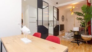 Una sala de estar moderna con una mesa de comedor de madera, sillas rojas, un sillón estampado, una mesita auxiliar, una lámpara y una planta grande en maceta.