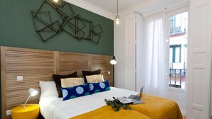 Un dormitorio con cabecera de madera, una cama con edredón blanco y dos otomanas amarillas, un ordenador portátil sobre la cama, y un adorno de pared con diseño geométrico.