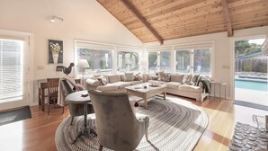 A spacious living room with a high wooden ceiling, large windows, a fireplace, and a pool view.