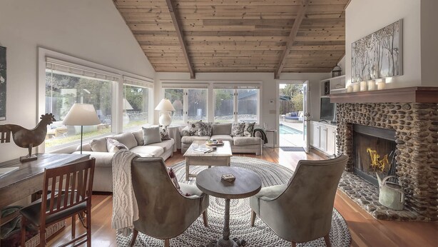 A spacious living room with a stone fireplace, large windows, and a wooden ceiling.