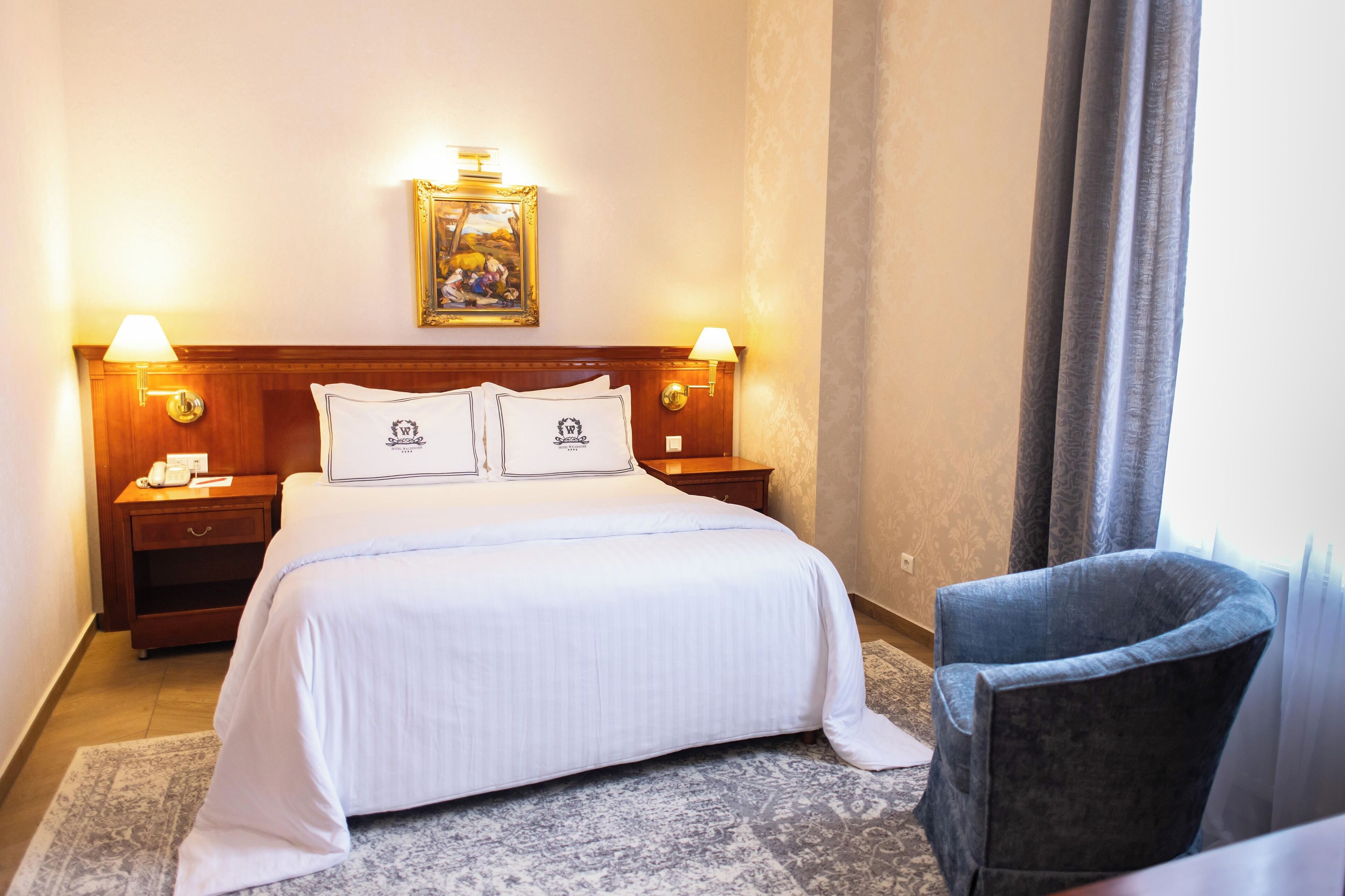 A hotel room with a large bed, two bedside lamps, a nightstand with a phone, a framed picture on the wall, and a chair by the window.