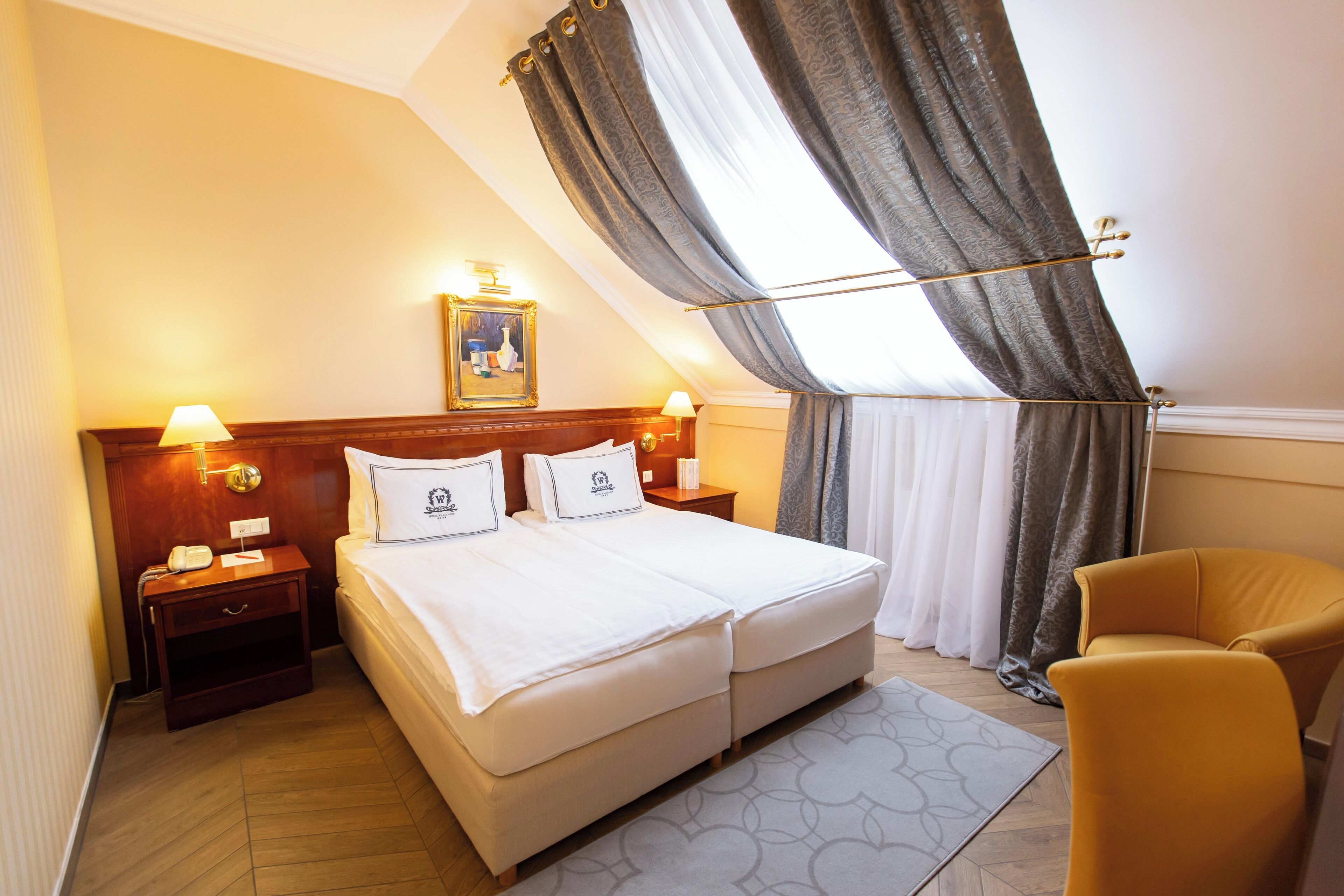 A hotel room with a double bed, two bedside tables with lamps, a nightstand, a chair, a painting on the wall, and a window with curtains.