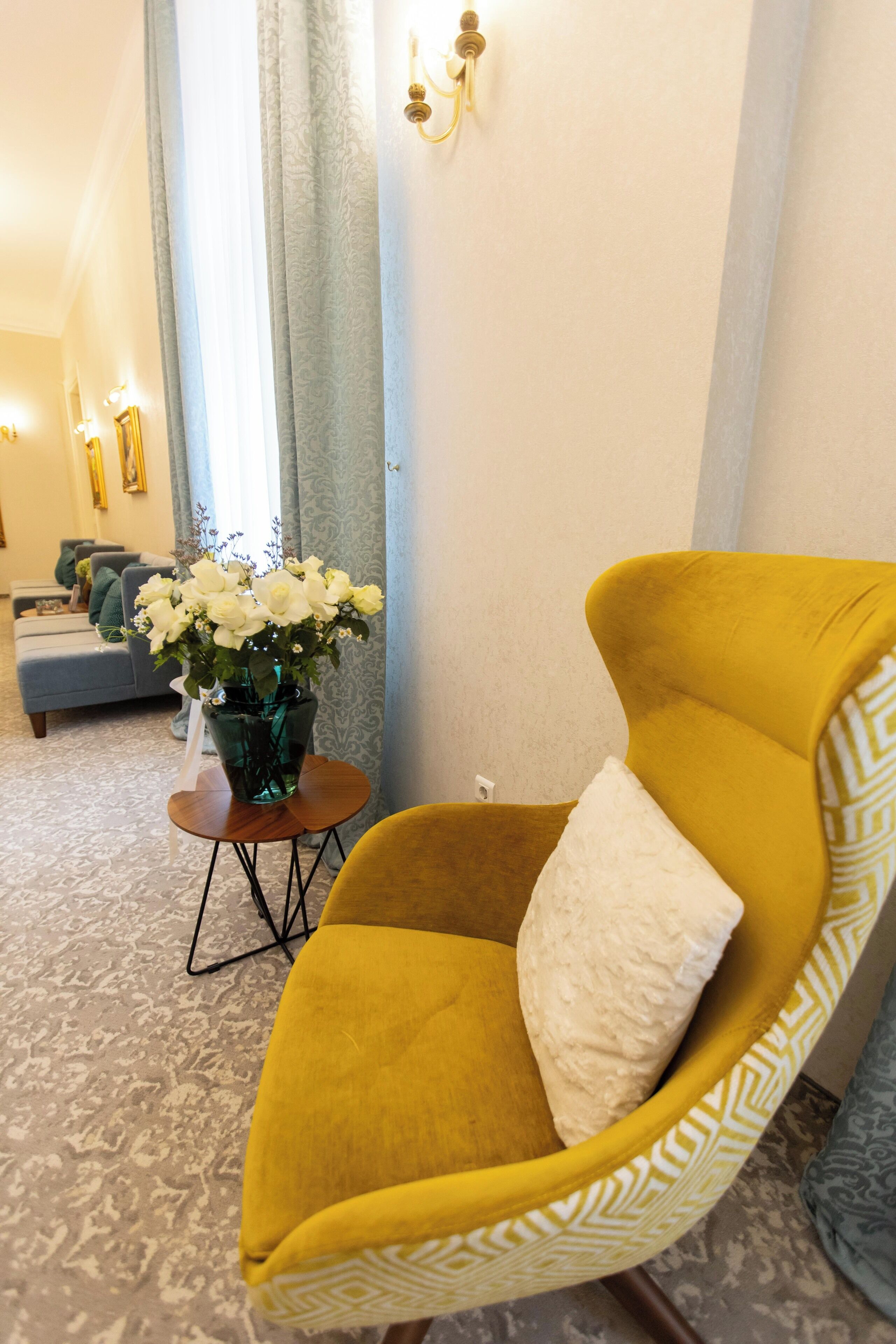A yellow armchair with a white cushion in a room with patterned carpet, a vase of white flowers, and a sofa in the background.