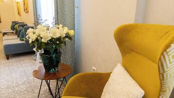 A yellow armchair with a white cushion in a room with patterned carpet, a vase of white flowers, and a sofa in the background.