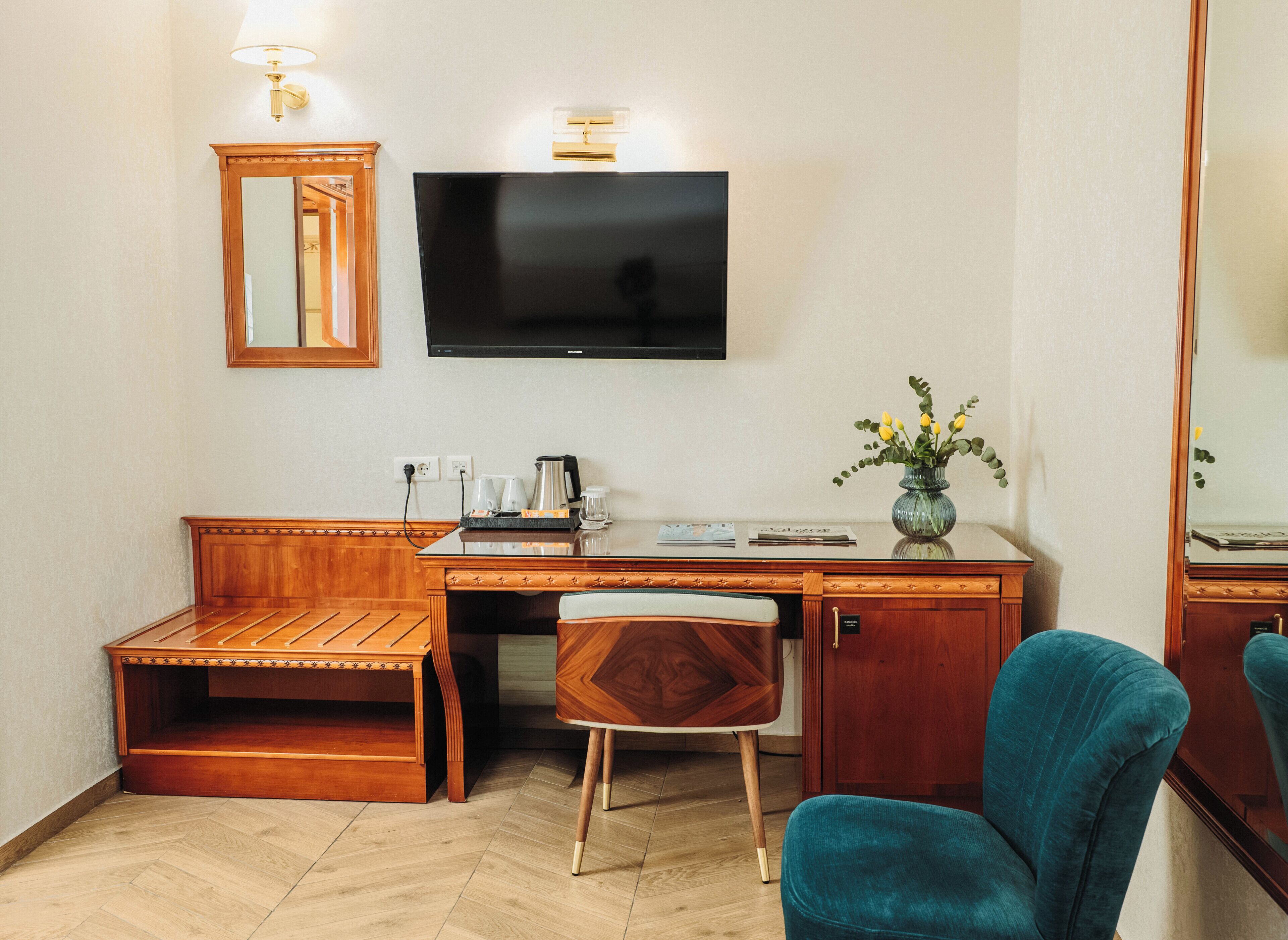 Habitación de hotel con escritorio de madera, televisor, espejo, un pequeño rincón de descanso y un jarrón con flores.