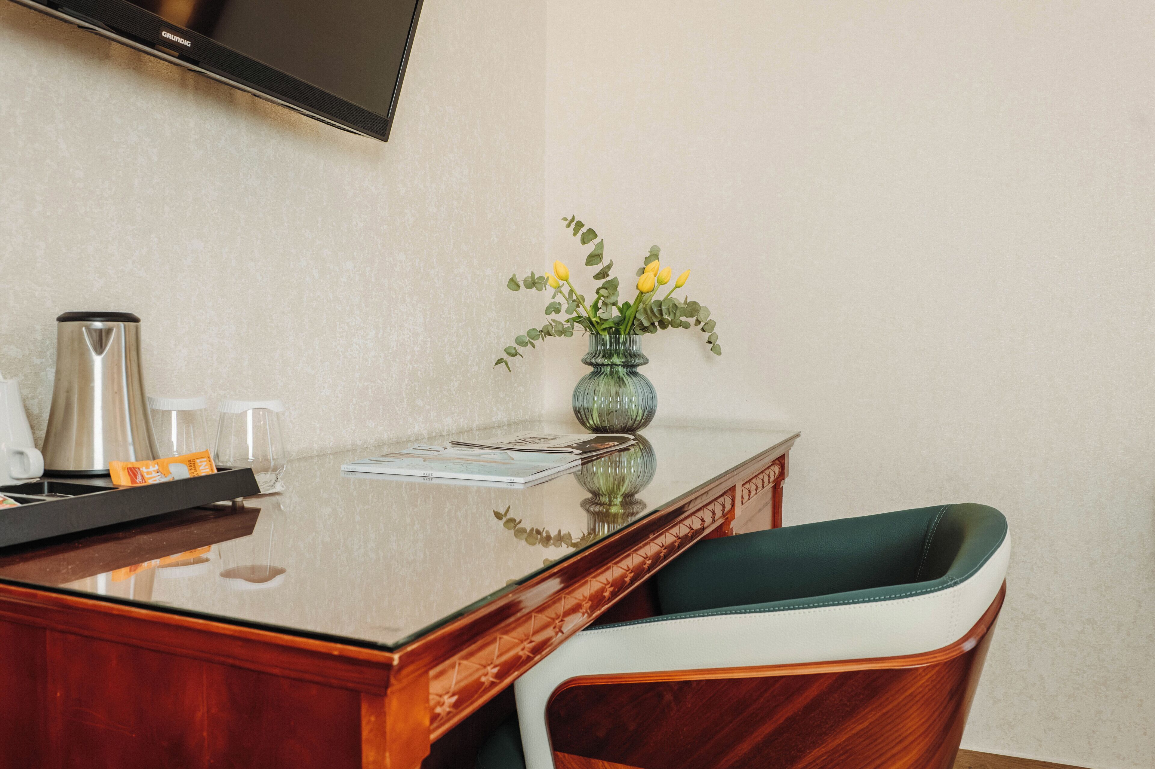 Una habitación de hotel con un escritorio de madera, un jarrón con flores, una cafetera y un televisor montado en la pared.