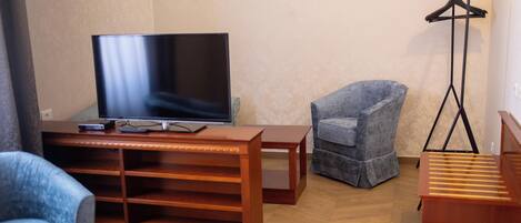 A living room with a flat-screen TV, a blue armchair, a wooden coffee table, and a floor lamp.