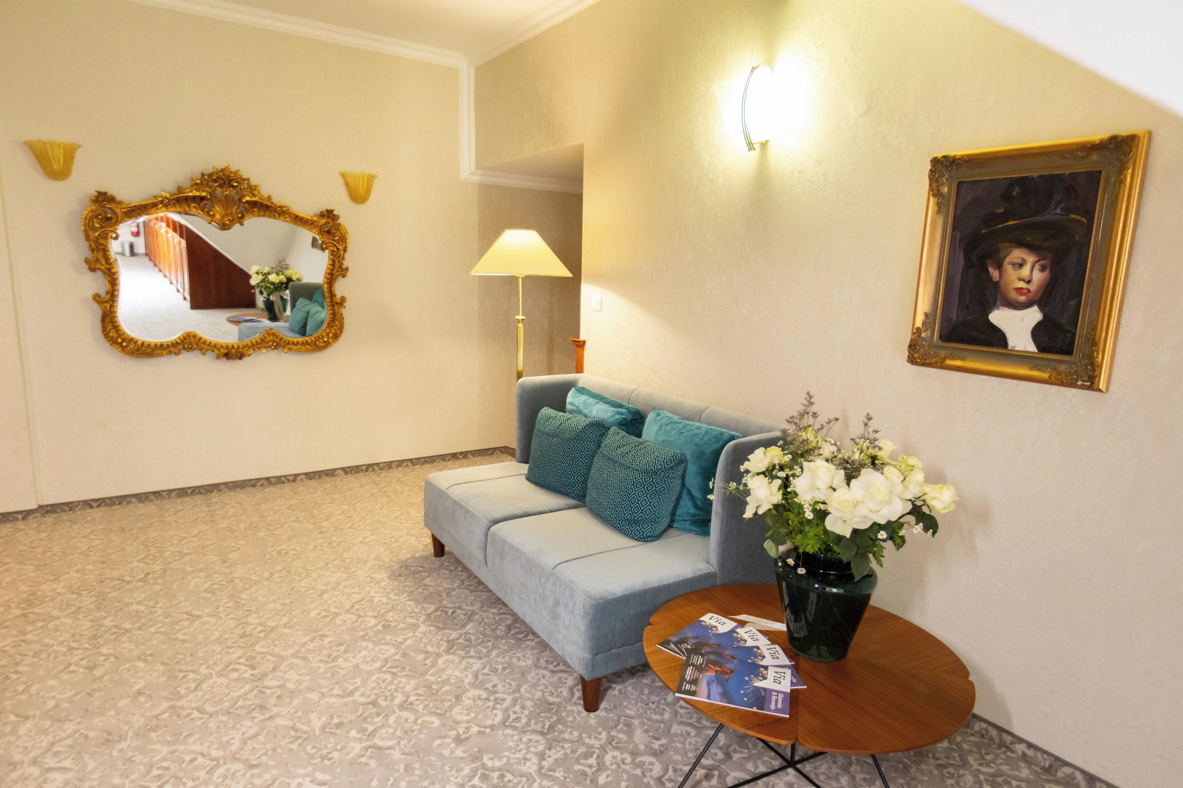 Un hall d’hôtel avec un canapé bleu clair, une table basse ronde en bois surmontée d’un vase de fleurs blanches, un miroir doté d’un cadre orné et un portrait encadré accroché au mur.