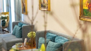 A living room with a grey sofa, a coffee table, and a vase of flowers. There are framed pictures on the wall and a lamp on the table. - Hotel Waldinger (Osijek)