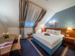 Une chambre d’hôtel avec un lit, un bureau, une chaise, une petite table ornée d’un vase de fleurs et une fenêtre avec des rideaux.
