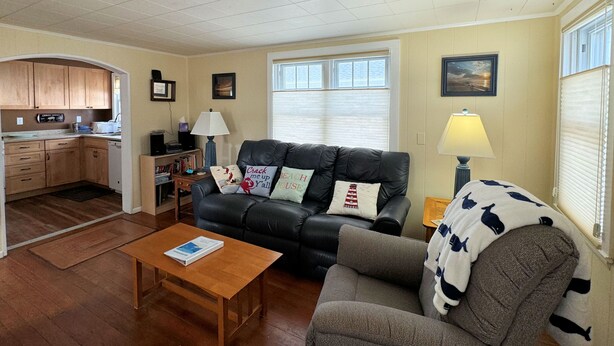 A living room with a black leather sofa, a grey armchair, a wooden coffee table, and a kitchen area with wooden cabinets and a window. - Sunshine House - 3BR home close to everything, screen porch! No Linens (Chincoteague)