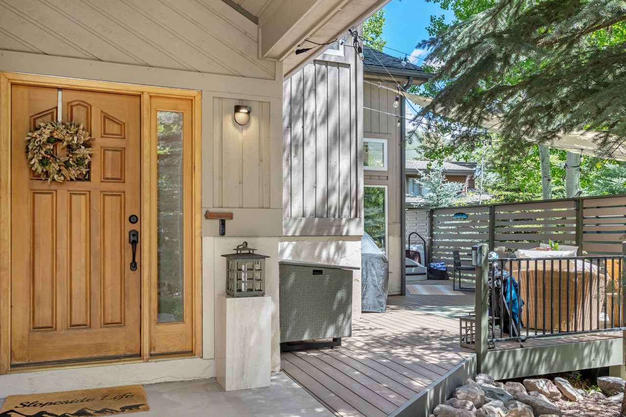 Une porte d’entrée en bois ornée d’une guirlande, dotée d’un panneau de verre et d’une lanterne, et un espace extérieur aménagé avec un coin salon couvert et un barbecue.