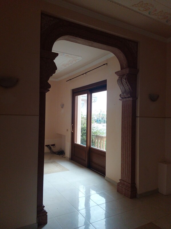 An interior corridor with a tiled floor, a wooden door with glass panels, and a view of greenery outside.