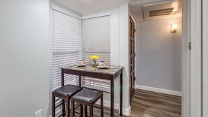 Un petit coin repas avec une table et deux chaises, une fenêtre avec des stores et une bouche d’aération fixée au mur.