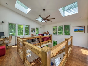 A spacious room with a high ceiling, skylights, a wooden bed with a red blanket, a desk with a computer, and large windows offering a view of trees. - Private Waterfalls, Creek, Fire Pits, Hot Tub, Private Hiking & Level 2 Charging (Asheville)