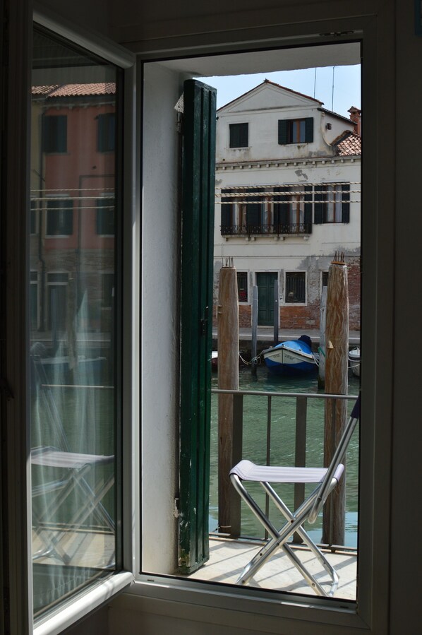 Vista a través de una ventana con una silla metálica afuera, un edificio con balcón y un barco en el agua.