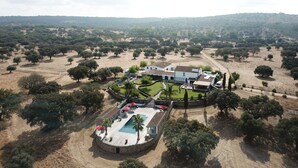 An aerial view of a property with a swimming pool, a large white building, and a surrounding landscape of trees and open fields.