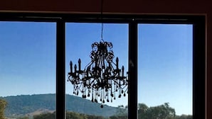 A room with a large window overlooking a dry landscape with trees and hills. A chandelier hangs from the ceiling, and there is a bench and a table with a vase of flowers in the foreground.