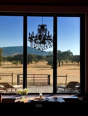 A room with a large window overlooking a dry landscape with trees and hills. A chandelier hangs from the ceiling, and there is a bench and a table with a vase of flowers in the foreground. - Splendid oasis in the heart of Alentejo "THE BARN" (Viana do Alentejo)