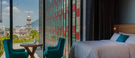 Habitación de hotel con un ventanal amplio que ofrece vistas de la ciudad, una cama con sábanas blancas y dos sillas color turquesa alrededor de una mesita pequeña.