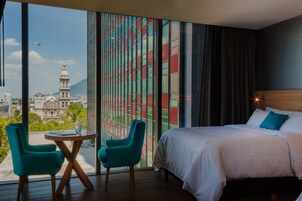 A hotel room with a large window offering a cityscape view, a bed with white linens, and two teal chairs around a small table.