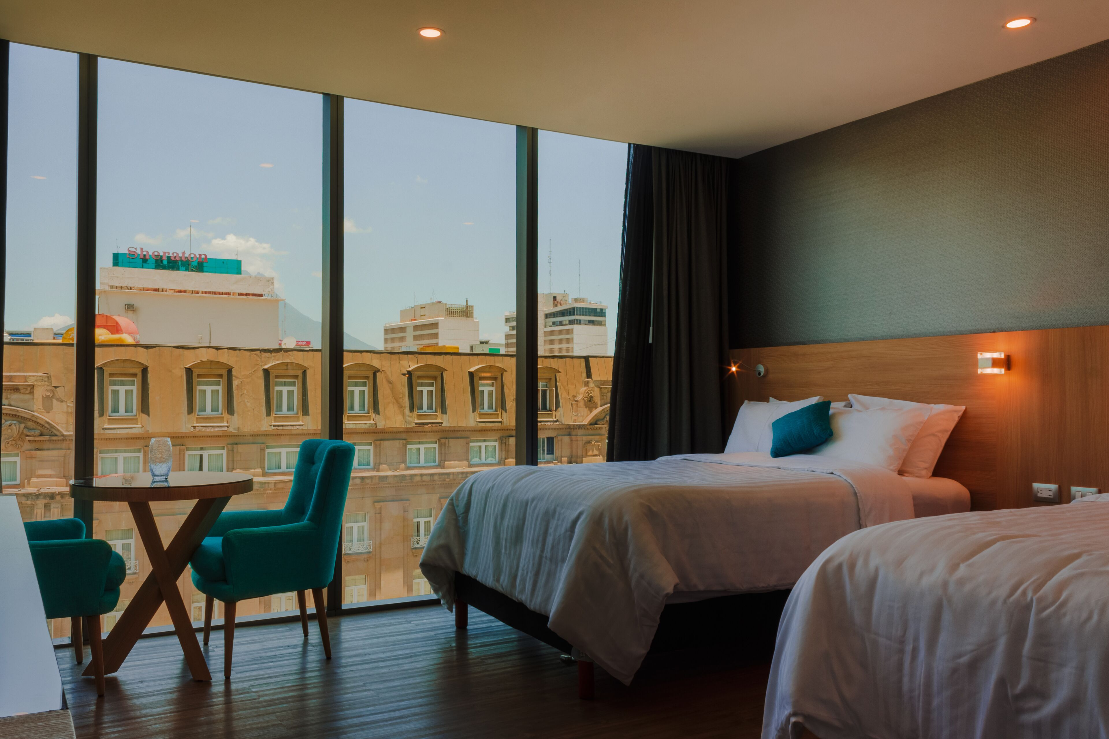A hotel room with a large window, two beds, a small table with chairs, and a view of the cityscape.