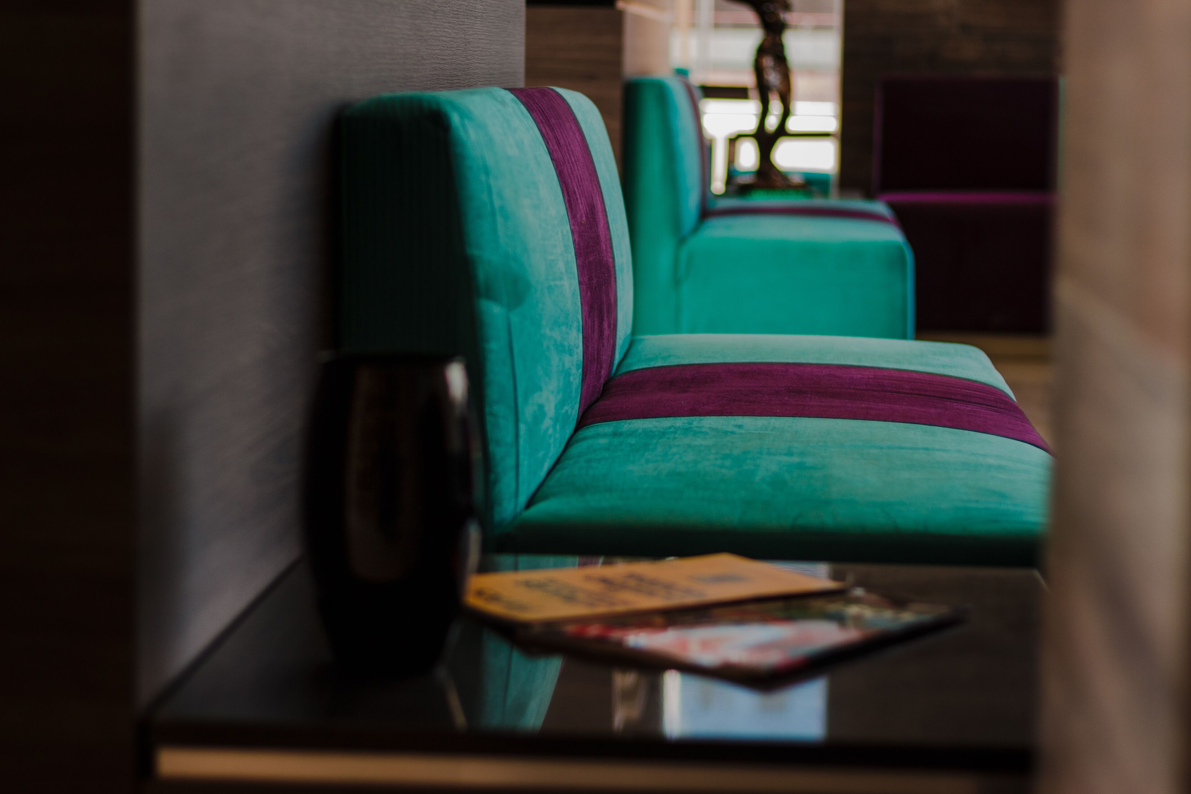 Ein blaugrünes Sofa mit violettem Streifen, eine dunkel gefärbte Vase und eine Zeitschrift auf einem schwarzen Tisch in einem Raum mit neutralen Wänden und violetten Akzenten.
