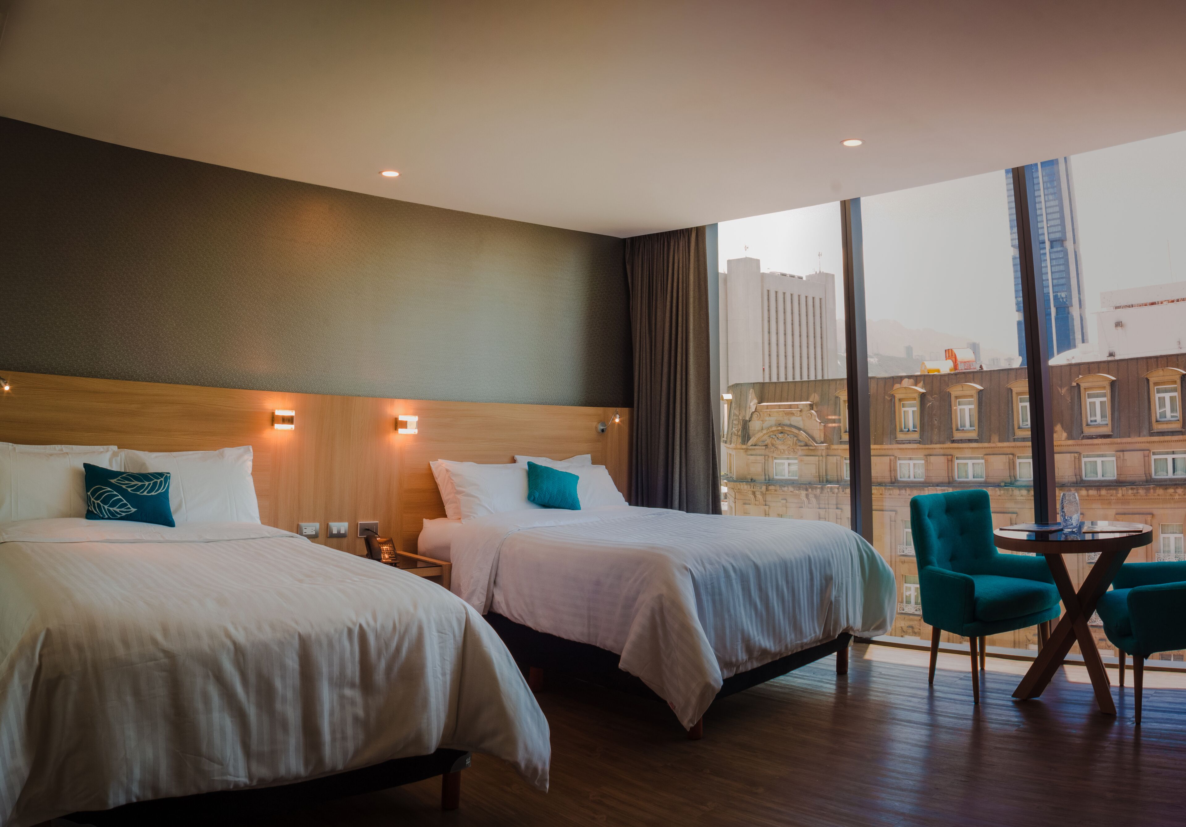 Quarto de hotel com duas camas, área de estar com mesa e cadeiras, e uma janela grande com vista para a cidade.
