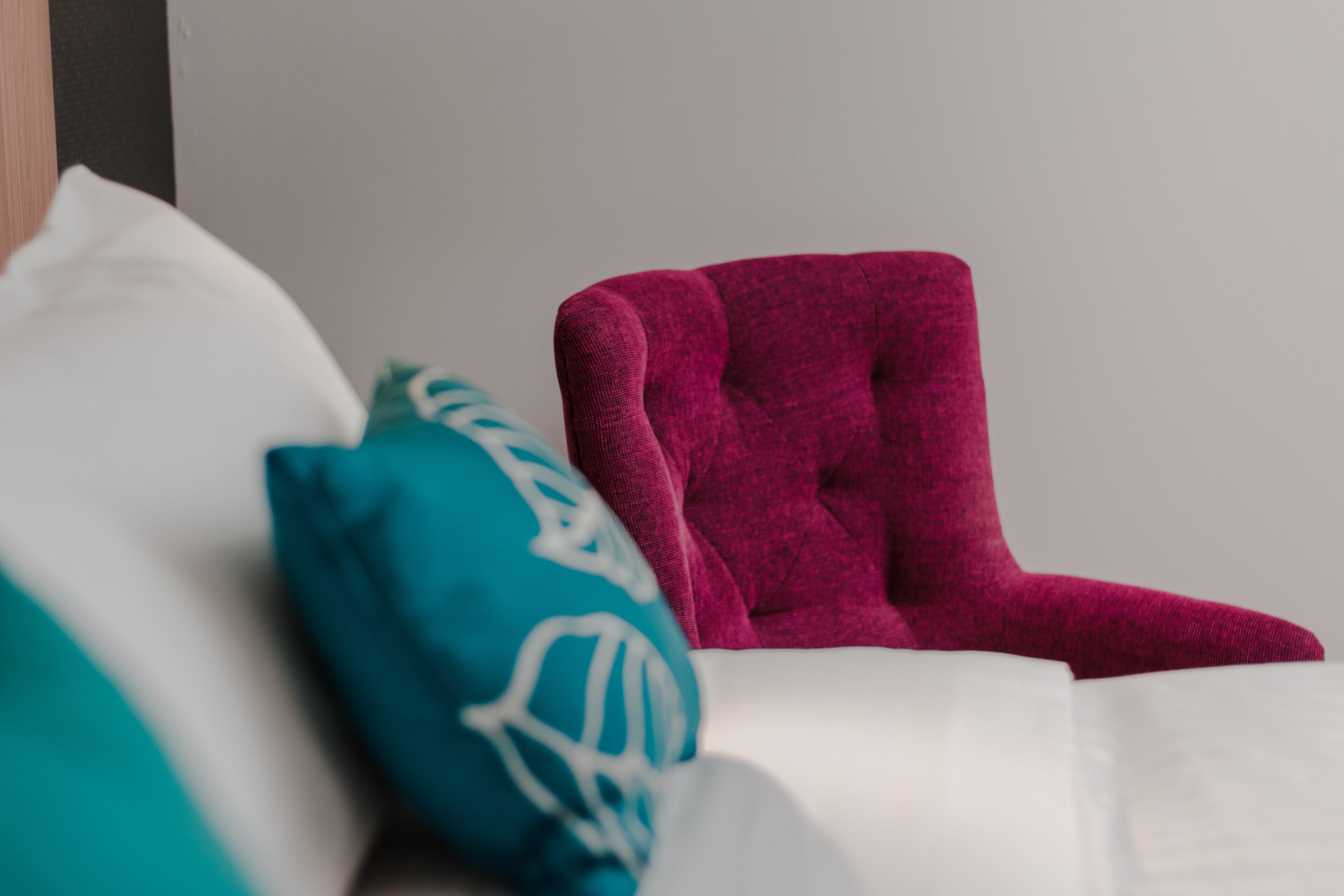 A bed with white bedding, a teal pillow, and a magenta chair.
