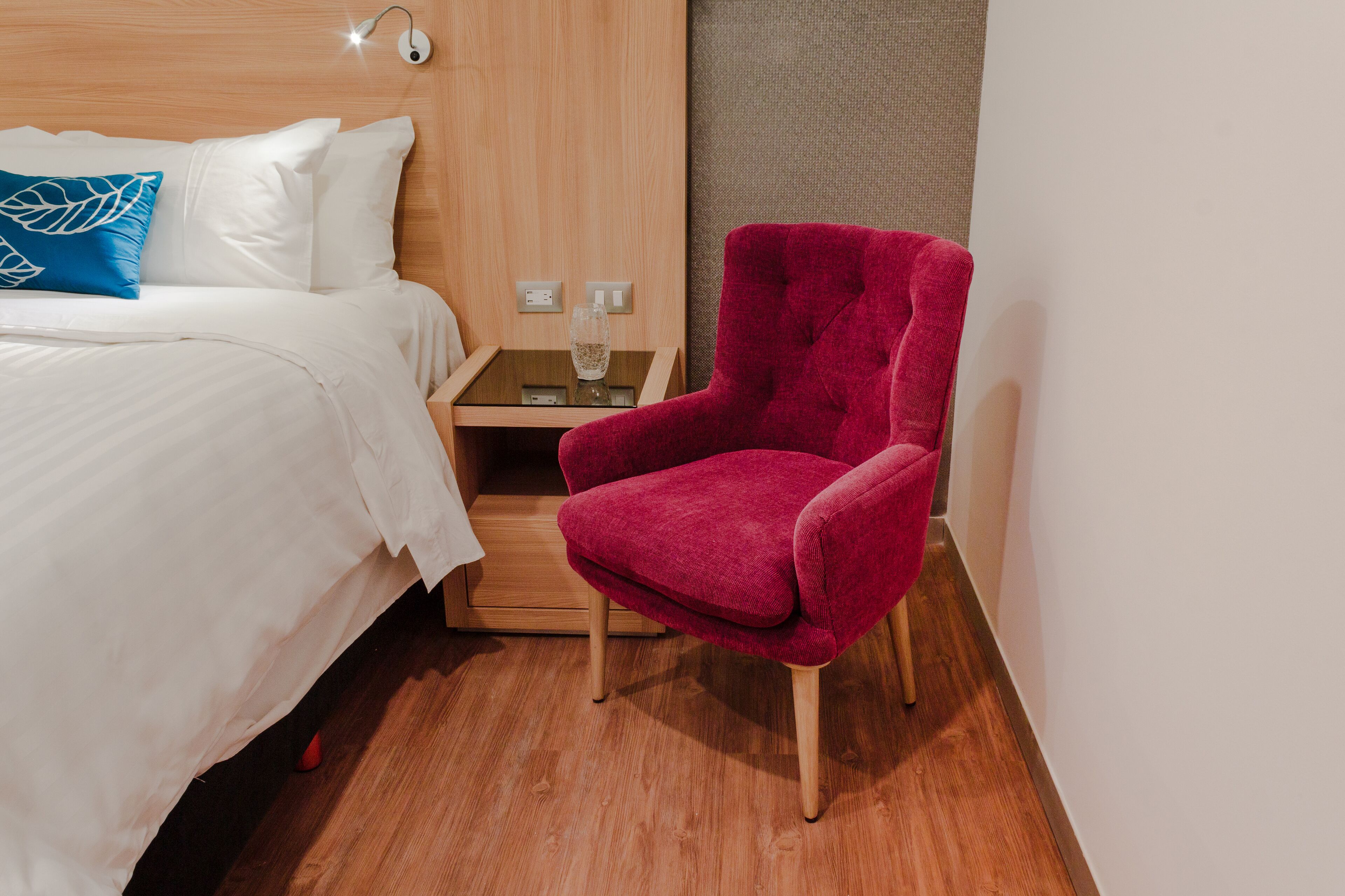 A hotel room with a bed, a red armchair, a wooden nightstand, and a glass of water.