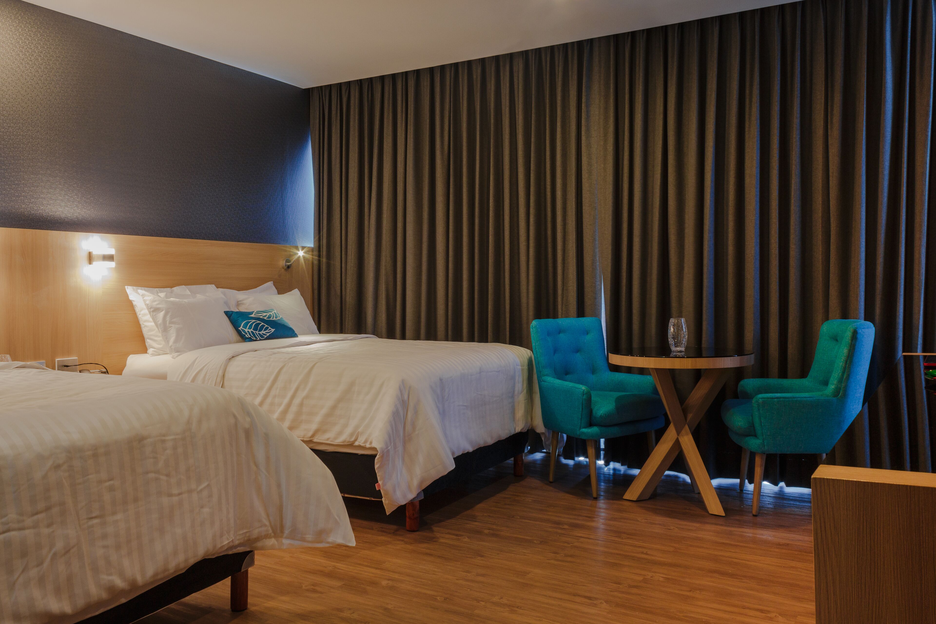 Une chambre d’hôtel avec deux lits, une tête de lit en bois, une grande fenêtre avec des rideaux, une table ronde avec deux chaises et un petit bureau en bois.