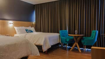 Une chambre d’hôtel avec deux lits, une tête de lit en bois, une grande fenêtre avec des rideaux, une table ronde avec deux chaises et un petit bureau en bois.