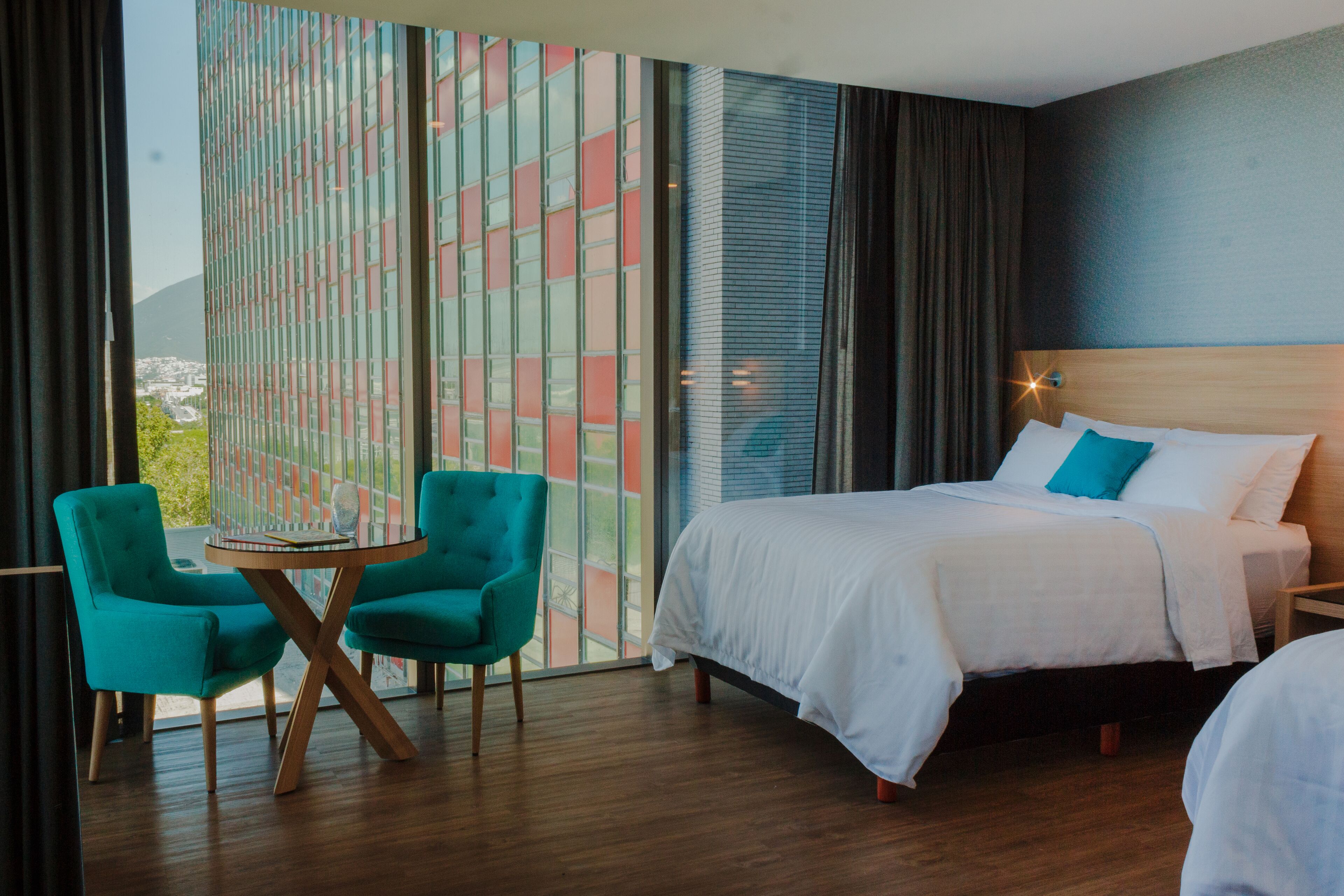 Hotelkamer met een bed, twee blauwgroene stoelen, een kleine tafel en een groot raam met uitzicht op gebouwen.