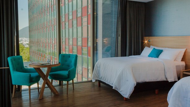 Habitación de hotel con cama, dos sillas color turquesa, una mesita y un ventanal con vistas a edificios.