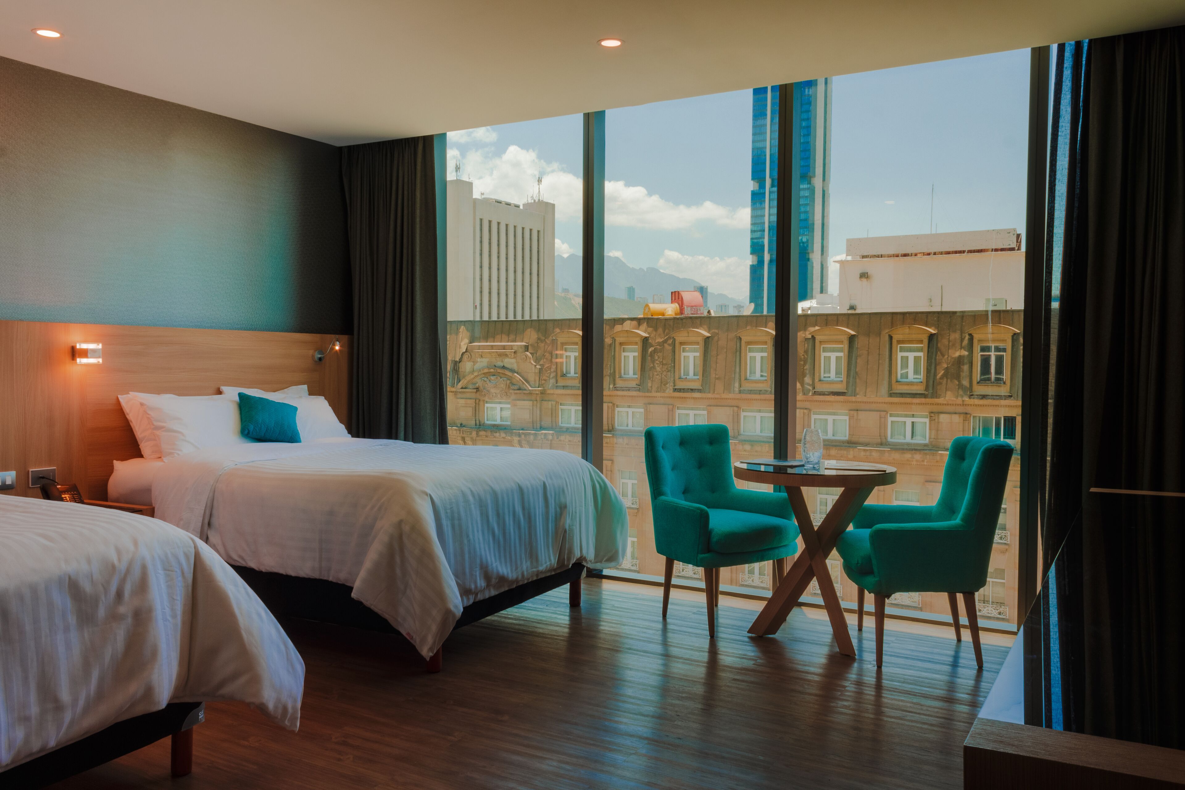 A hotel room with two beds, a small table, two chairs, and large windows offering a city view.