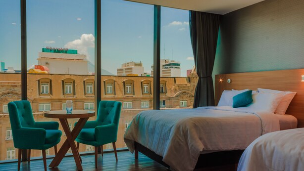 Una habitación de hotel con un ventanal grande, dos camas, una mesa redonda y dos sillas color turquesa.