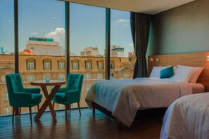 A hotel room with a large window, two beds, a round table, and two teal chairs. - Hotel Kavia Monterrey (Monterrey)