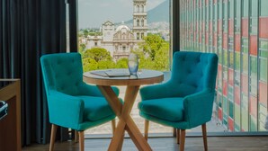 Una habitación con un ventanal grande, dos sillas color turquesa, una pequeña mesa redonda y vistas a un edificio histórico.