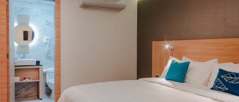 Habitación de hotel con cabecera de madera, cama con ropa blanca y dos almohadas azules, espejo redondo con luz, y baño pequeño con piso de mármol.