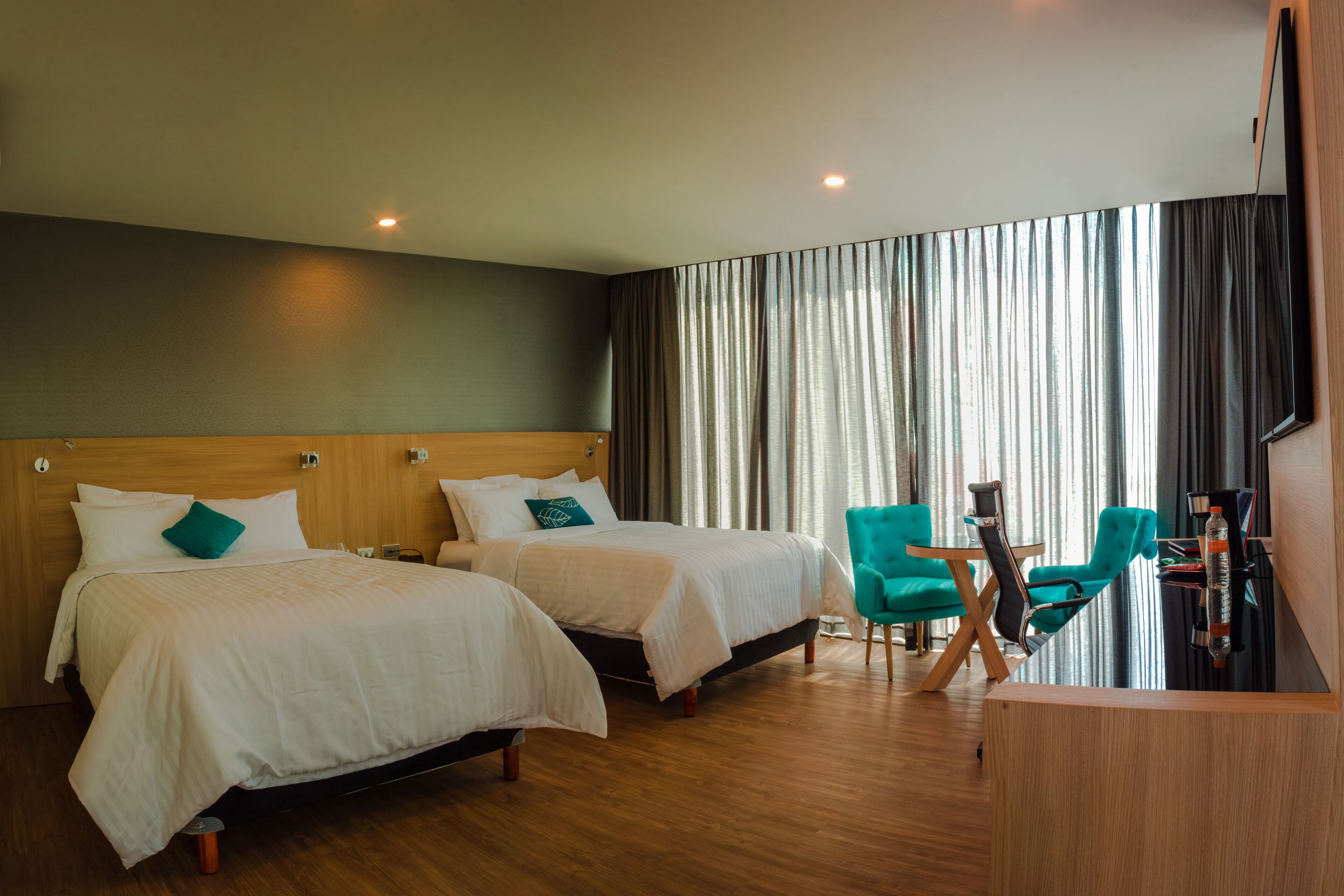 A hotel room with two beds, a wooden headboard, a desk with a chair, and a large window with curtains.