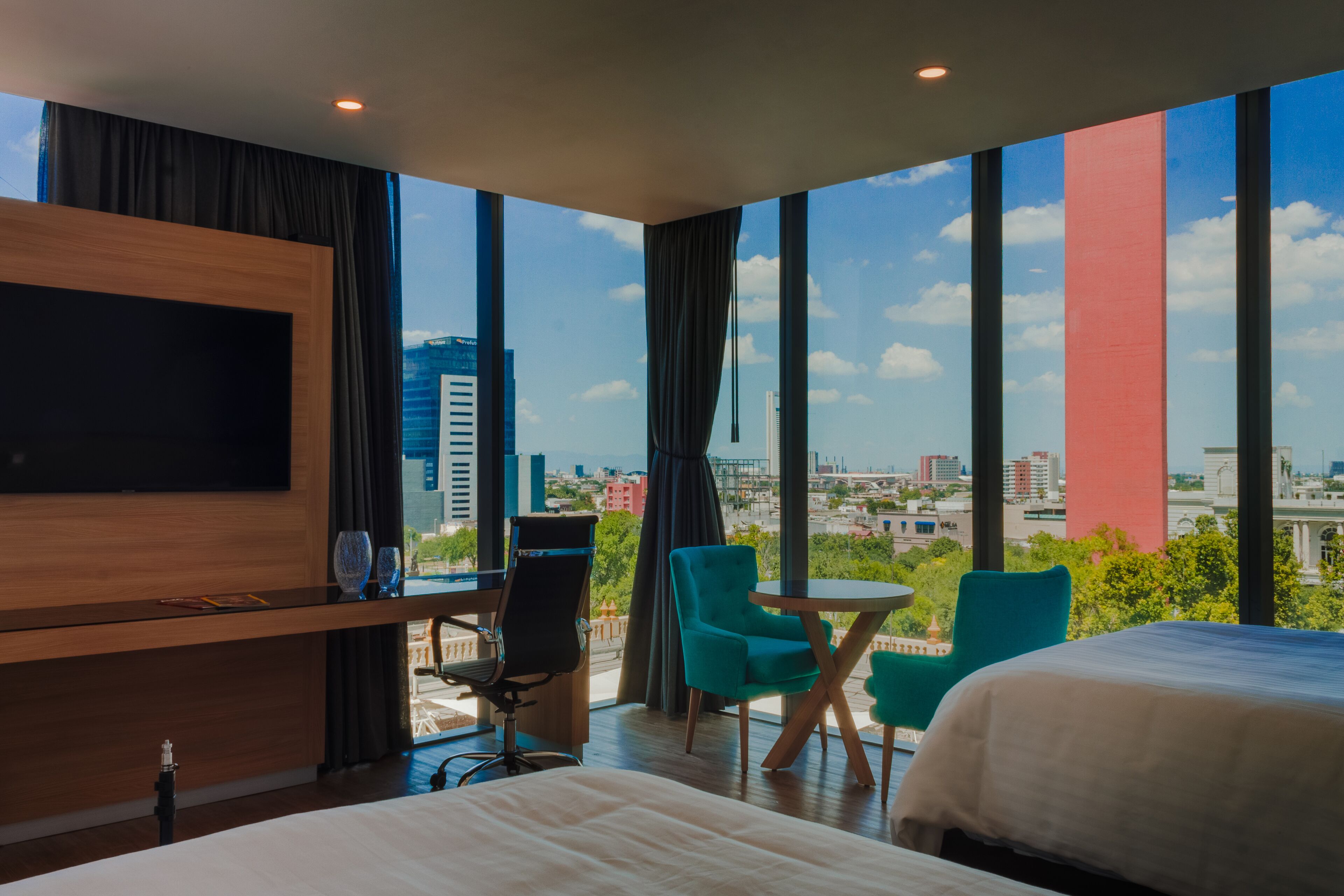 A modern hotel room with a large TV, a bed, two chairs, a small table, and a city view through large windows.