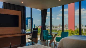 Habitación de hotel moderna con una televisión grande, una cama, dos sillas, una mesita y vistas a la ciudad a través de amplios ventanales.