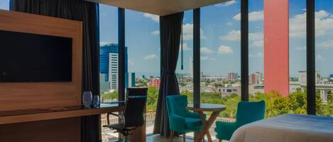 A modern hotel room with a large TV, a bed, two chairs, a small table, and a city view through large windows.