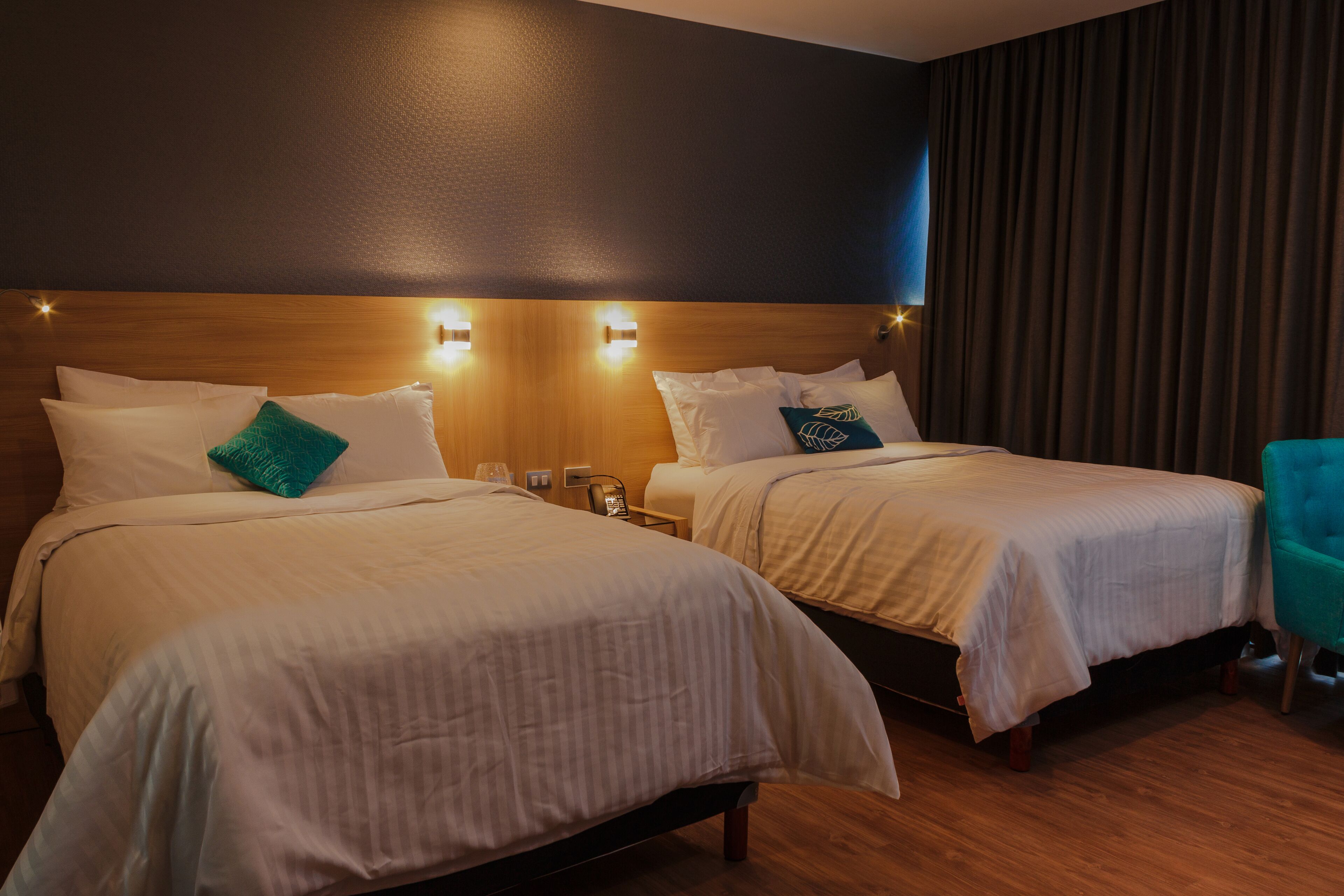Habitación de hotel con dos camas, cabeceros de madera y una silla verde azulada.
