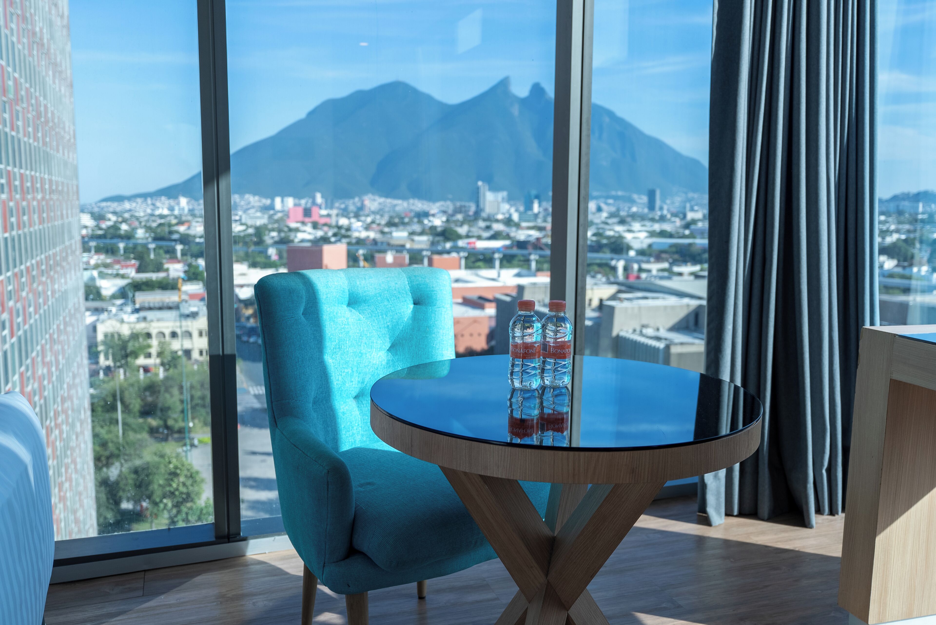 Une pièce dotée d’une grande fenêtre donnant sur le paysage urbain, une table ronde en verre avec une bouteille, et un fauteuil bleu.