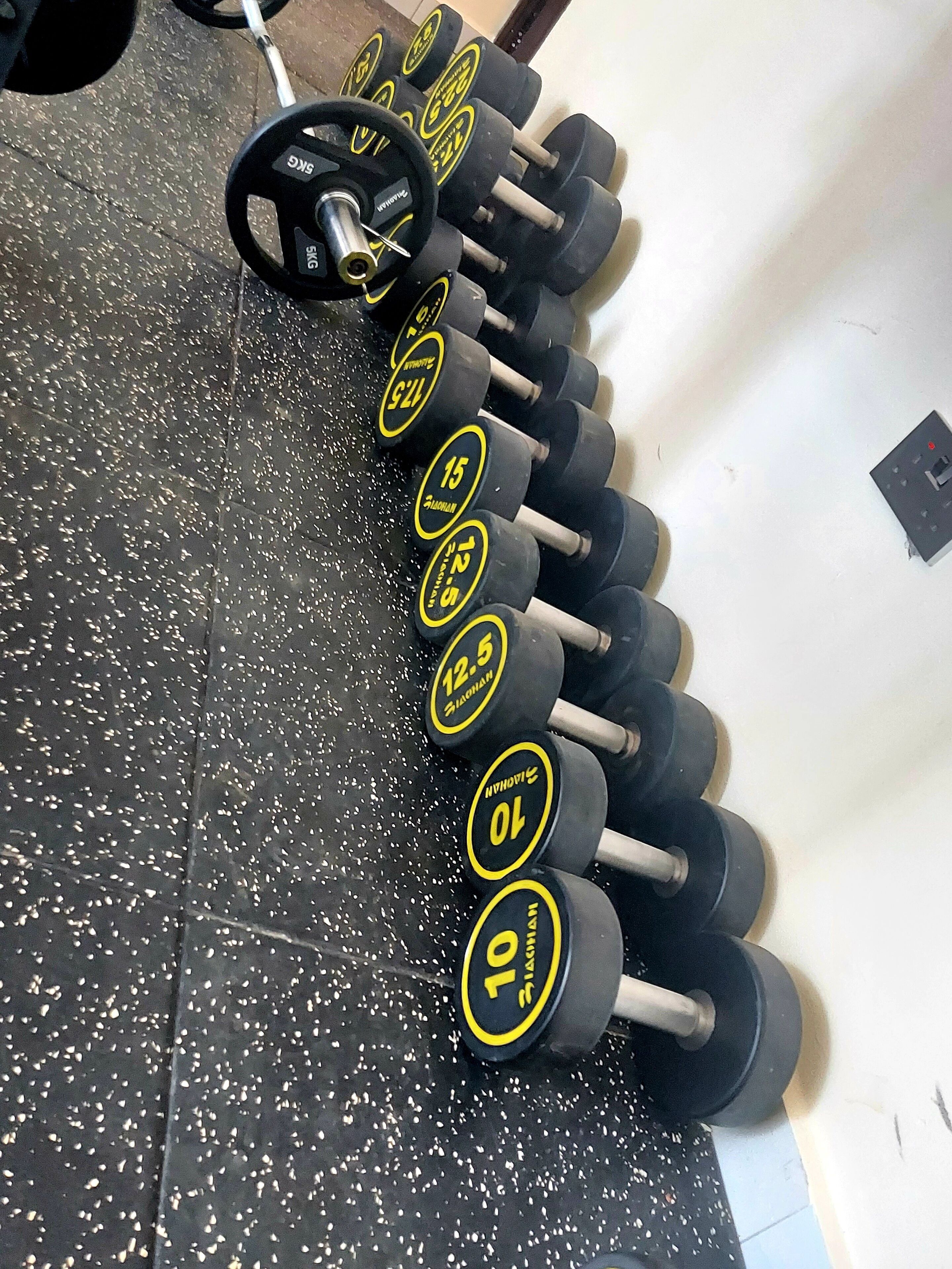 Una hilera de mancuernas con pesas que van de 10 a 15 kilogramos, dispuestas sobre un suelo negro moteado en un gimnasio.