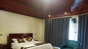 A hotel room with a wooden ceiling, a bed with white bedding and patterned pillows, a teal armchair, a small round table, and a ceiling fan. - Golden View Hotel Ibanda (Ibanda)