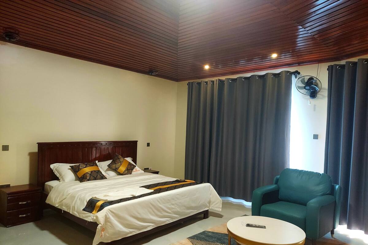 A hotel room with a wooden ceiling, a bed with white bedding and patterned pillows, a teal armchair, a small round table, and a ceiling fan.